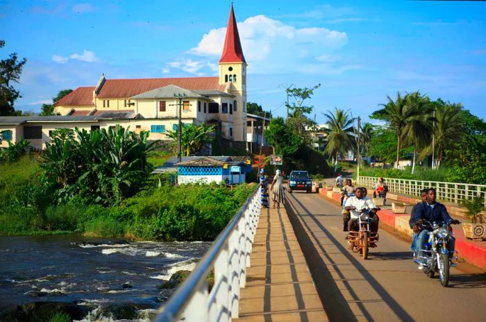 Motorcyclists traverse a bridge in a quaint town, with a church in view.