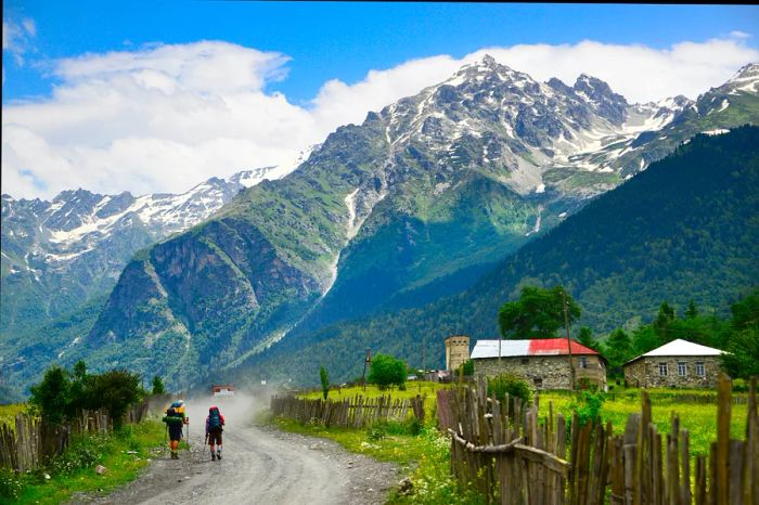 Two hikers traverse a path leading towards a mountain peak, passing a small settlement