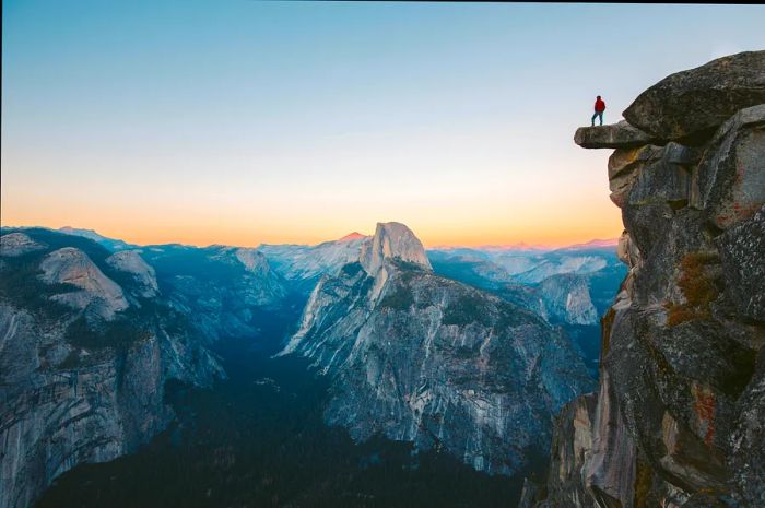 A hiker stands on a jutting rock, soaking in the breathtaking view at Glacier Point during the evening.