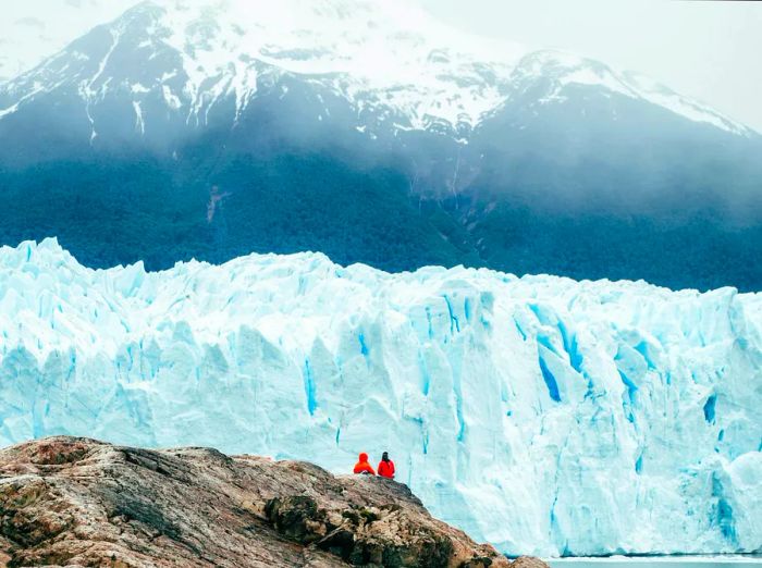 Two individuals enjoy the view from a lookout, captivated by a towering blue-white ice wall before them