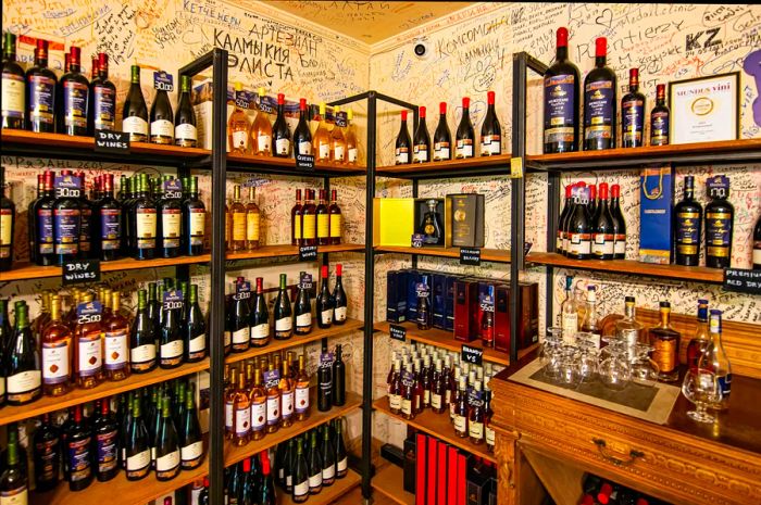 A wine shop displays shelves brimming with various types of Georgian wine, while the walls behind them feature vibrant graffiti-style writing.