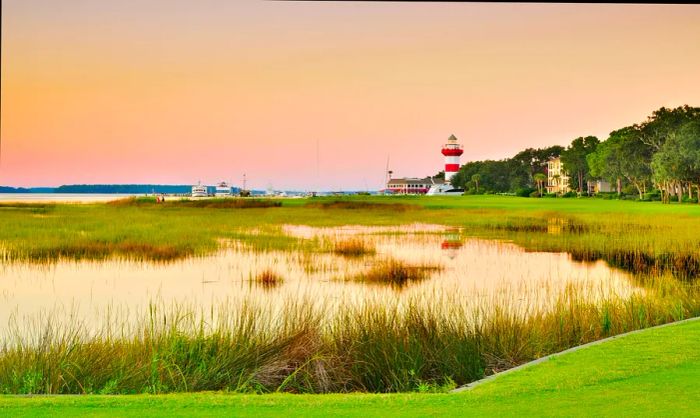 A view of a golf green adjacent to the water, with reeds bordering the harbor and a distant red-and-white lighthouse.