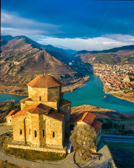 An ancient monastery perched on a hill overlooks the valley where two rivers converge, with a town visible on the opposite riverbank.