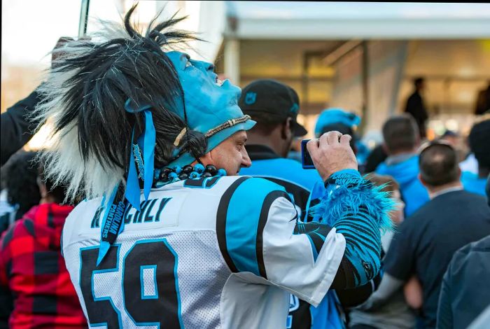 A close-up of a Carolina Panthers fan, wearing a jersey and a mask pushed back on his head, capturing a moment with his camera