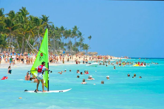 A windsurfer sets out on the ocean just offshore, with many people enjoying the sea.