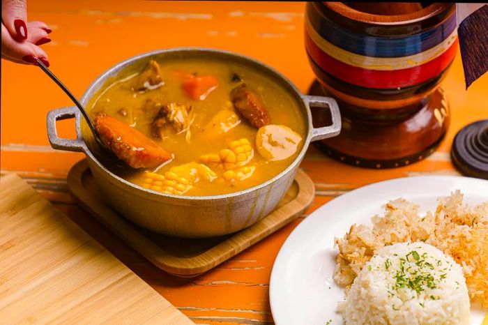 A hearty bowl of meat stew featuring large chunks of corn, accompanied by rice