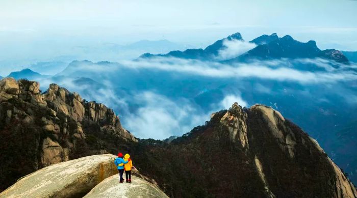 Two hikers admire the view from a rocky peak, with clouds shrouding the mountains in the background.