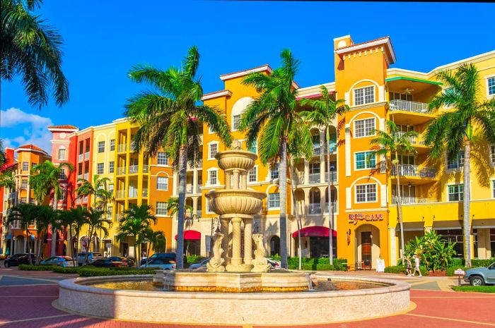 A fountain surrounded by palm trees stands in front of a row of tall, colorful buildings reminiscent of European architecture.