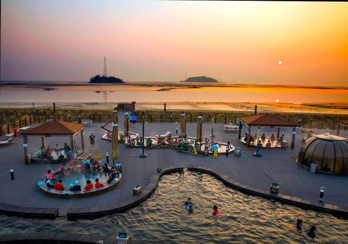 An aerial view of a pool area featuring several smaller hot-water pools in a coastal spa, with a sunset in the background