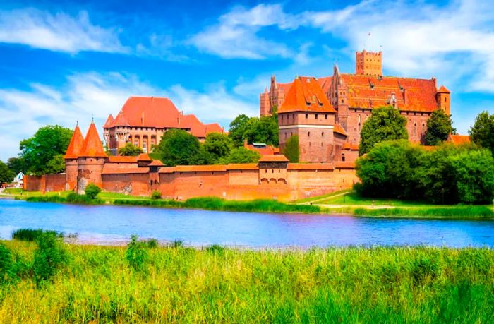 A massive red-brick castle adorned with numerous towers and battlements rises along the serene riverbank.