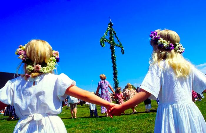 Two girls in white dresses and floral crowns join hands as they twirl around a maypole, surrounded by others holding hands. In Sweden, the Summer Solstice is celebrated as Midsummer.