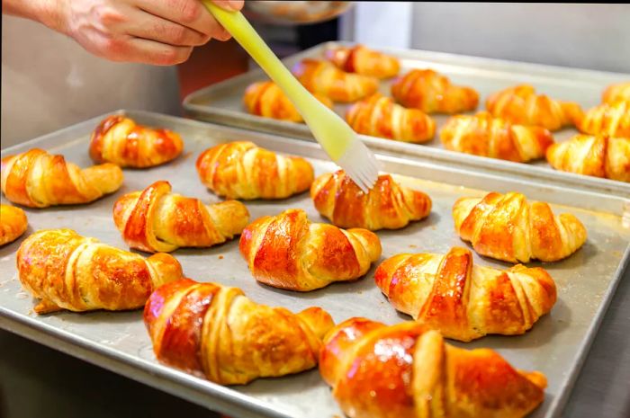 A pastry chef is applying a golden glaze to freshly baked croissants arranged on trays in a professional bakery.