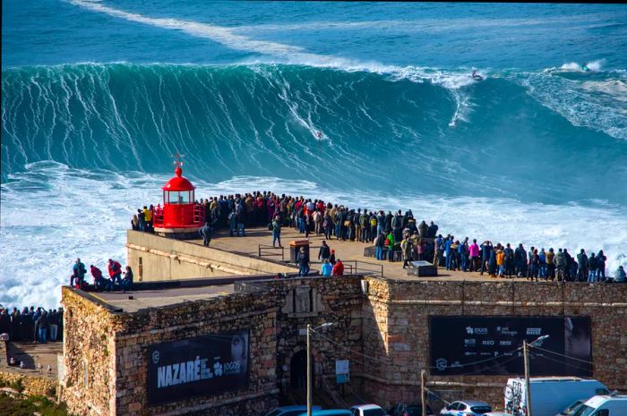 Onlookers gather along a walkway to a lighthouse, eager to watch the massive waves being surfed close to shore.