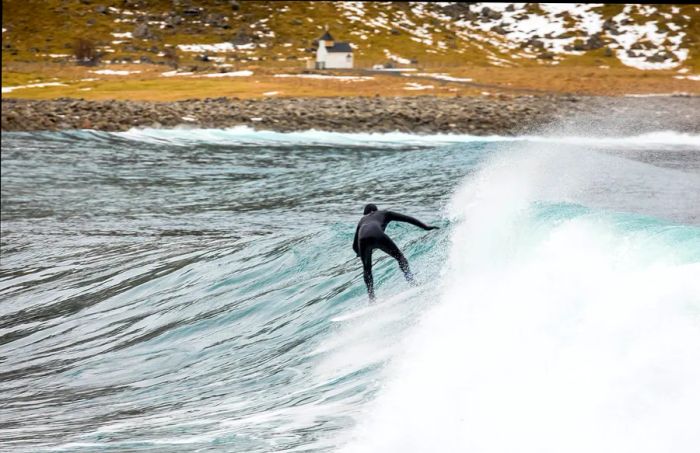 A fully geared surfer rides a wave at a bay, with snow capped rocks lining the beach.