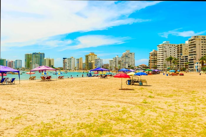 A beachfront area lined with apartment buildings, featuring numerous loungers and shaded seating under gazebos.