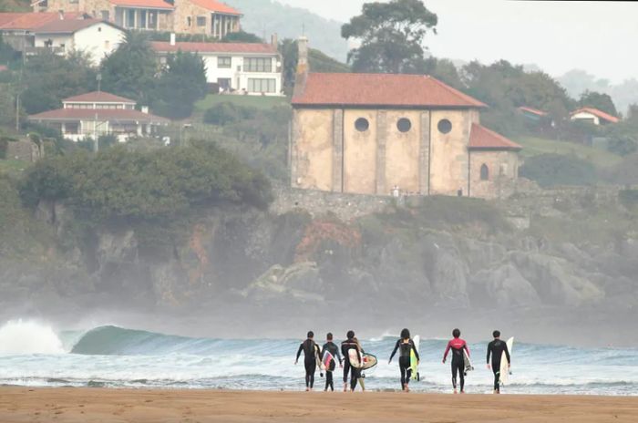 A group of surfers walk purposefully towards the waves, boards in hand, with a large church and homes visible on the hillside in the background.