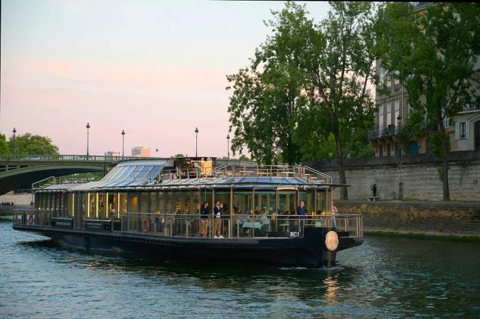 A group of guests enjoys the view from the deck of the Ducasse sur Seine, a floating restaurant, as it elegantly cruises along the River Seine at twilight.