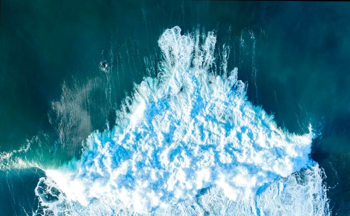 An overhead view captures the wild surf with a lone, daring surfer venturing near its edge.