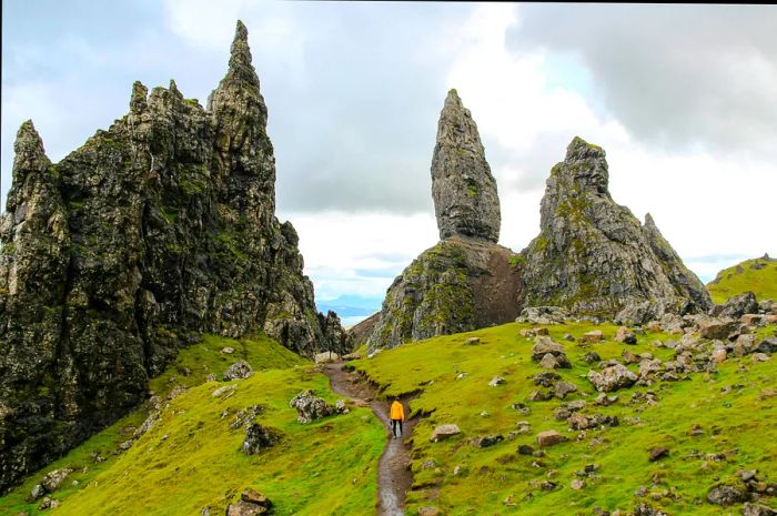 A girl in a yellow raincoat hikes along a trail towards The Old Man of Storr amidst unpredictable weather.