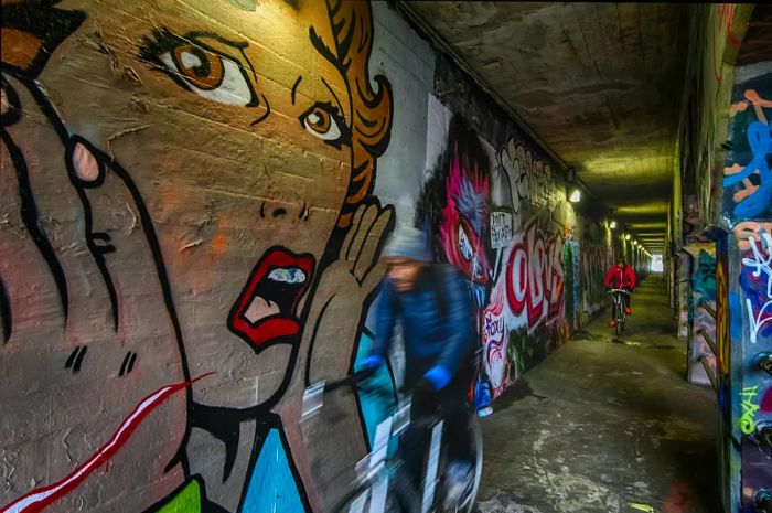 A striking mural of a startled woman, inspired by Roy Lichtenstein, adorns the wall of a graffiti-filled tunnel, as cyclists pass by.