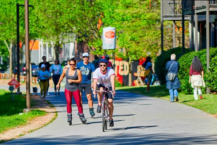 Various individuals are seen cycling, rollerblading, and walking along a section of the Atlanta BeltLine on a sunny day.