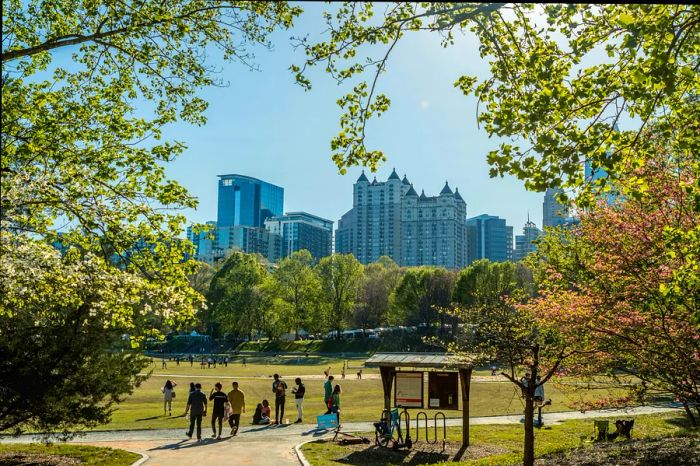 People are enjoying the expansive green spaces of Piedmont Park on a sunny day, with a backdrop of the Atlanta skyline visible through the trees.