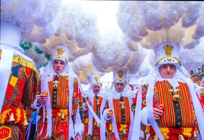 Individuals dressed in intricate costumes that envelop their bodies and heads gather in a hazy atmosphere for a carnival parade.