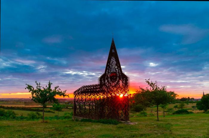 A sculpture composed of slats forming a chapel-like structure, illuminated by the setting sun streaming through it.