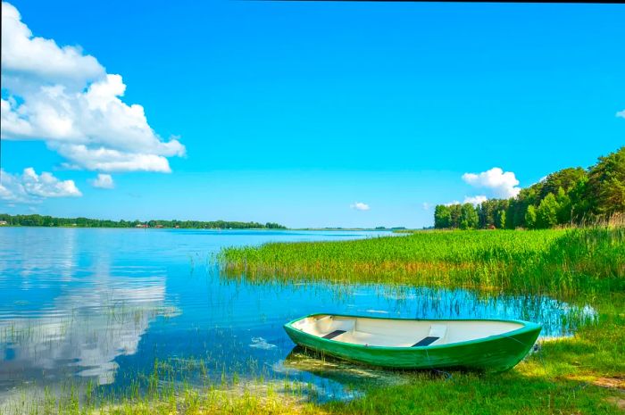 A small green rowing boat is docked at the edge of a vast lake on a sunny day.