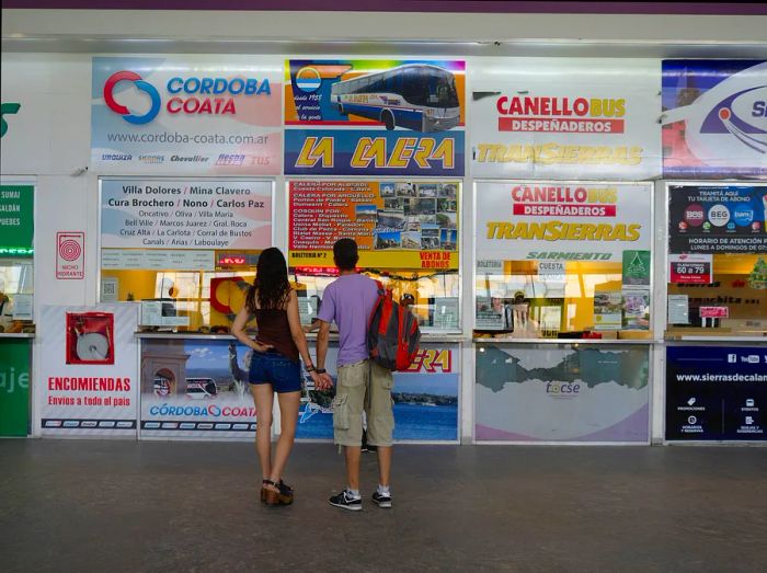 A couple gazes up at the bus station ticket office displays, hand in hand.