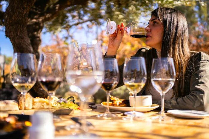 A woman at a wine-tasting event enjoys sipping red wine from a selection of white and red wines arranged on a table before her.