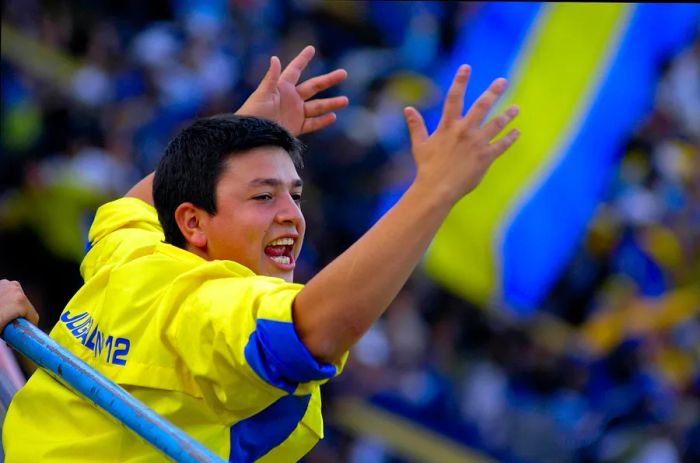 A teenager proudly sporting the yellow and blue of Boca Juniors raises his arms in celebration at a soccer match in Buenos Aires, Argentina.