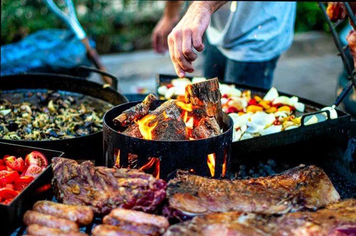 A man is tending to a large, wood-fired barbecue grill loaded with an array of meats and vegetables.