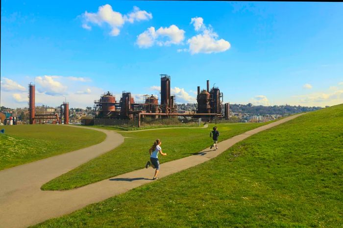 Two young children joyfully run along a sunny park path with the rusty remnants of old gas works nearby.