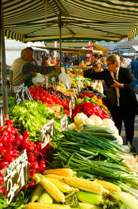 A woman is purchasing fresh produce at a market stall brimming with a wide variety of vegetables, including corn, radishes, and lettuce.