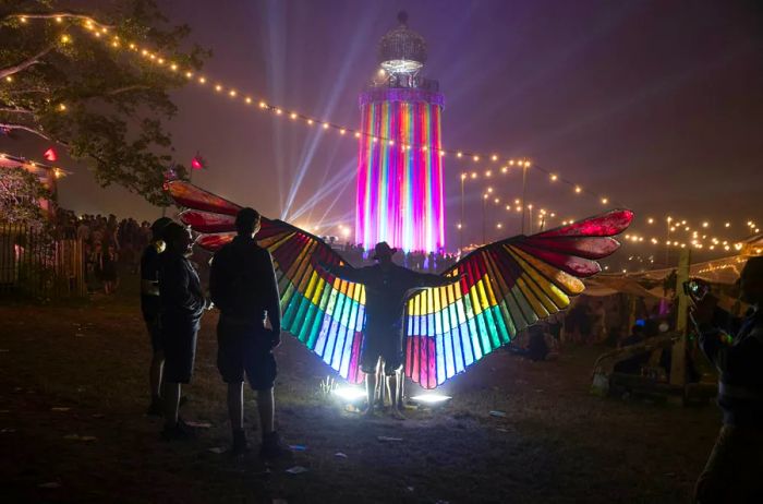 A person sporting massive rainbow-colored wings illuminated by a vibrant ride in the background