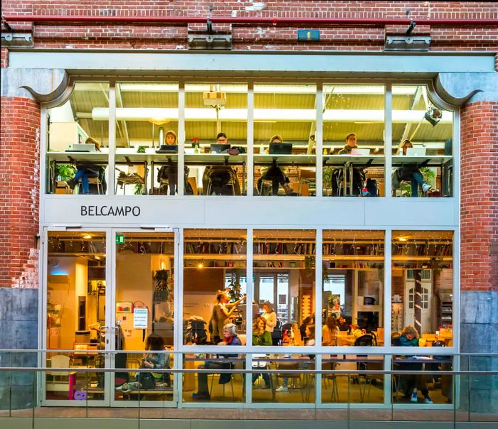 A spacious array of windows in a repurposed industrial building showcases people working at laptops and gathered at tables.