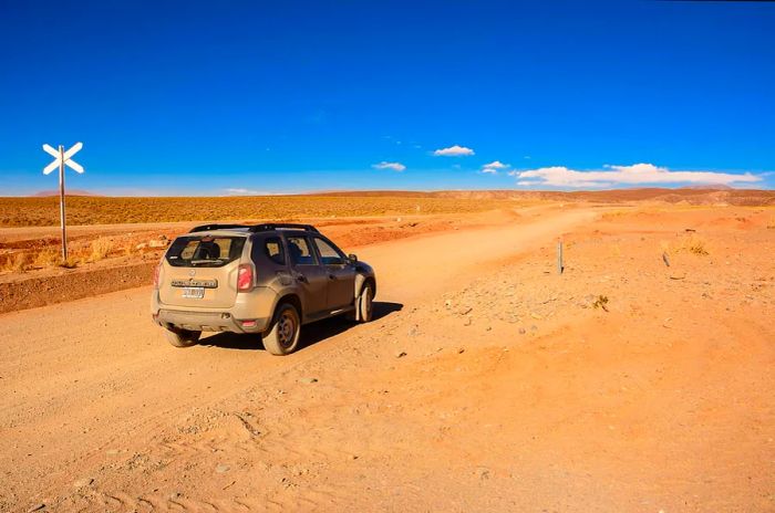 A dusty vehicle rests on an unpaved road, surrounded by the red sands of a desert landscape.