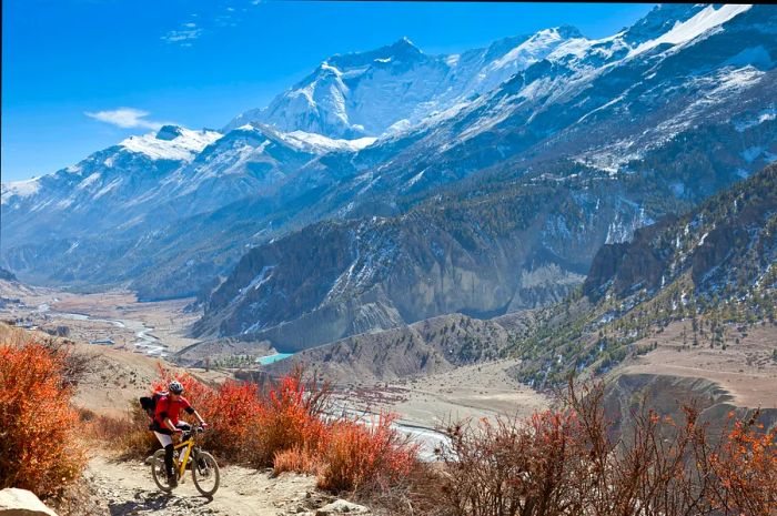 A male mountain biker has departed the village of Manang and is ascending towards Thorong La, surrounded by stunning scenery on the Annapurna Circuit, with Annapurna III and Gangapurna peaks visible on the right.