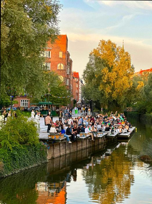 People relax on a deck with drinks beside a canal flowing through a town