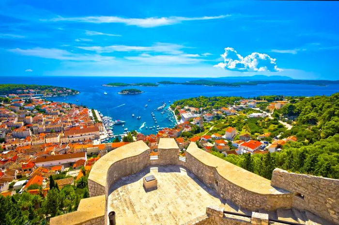 Fort battlements in the foreground reveal a scenic view of the harbor, the town's red rooftops, and islands in the distance.