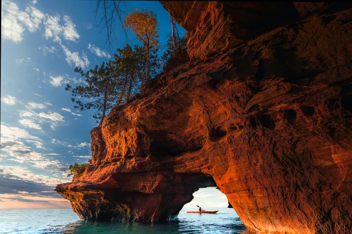 A kayaker framed by a red rock sea arch topped with trees