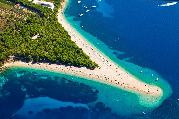 An aerial view of a peninsula adorned with trees and bordered by white-pebble beaches on both sides.