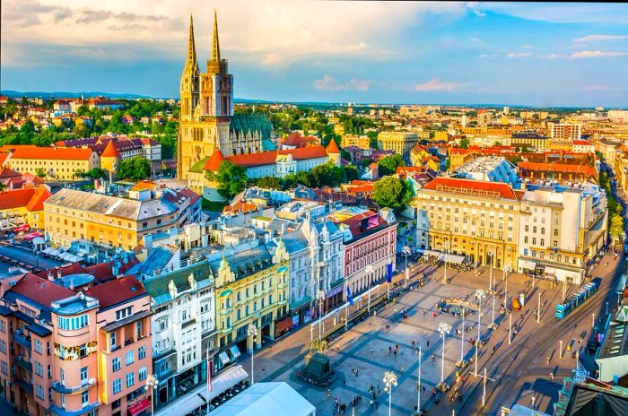 A bustling city square bordered by pastel-hued buildings and a skyline highlighted by a Gothic-style cathedral