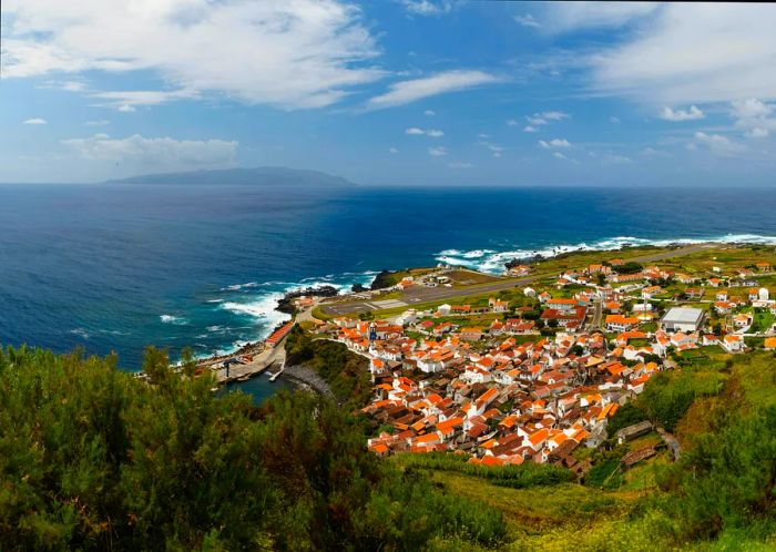 A sweeping view of the quaint village of Villa do Corvo, situated on Corvo Island. This photo captures a clear summer morning from one of the nearby viewpoints, showcasing the village's airport runway and harbor, along with the distant Flores Island.