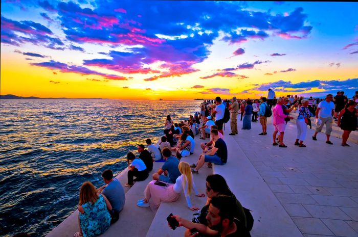 As the sun sets, people gather on the steps by the sea, watching as the sky transforms into a canvas of oranges, yellows, blues, and purples.