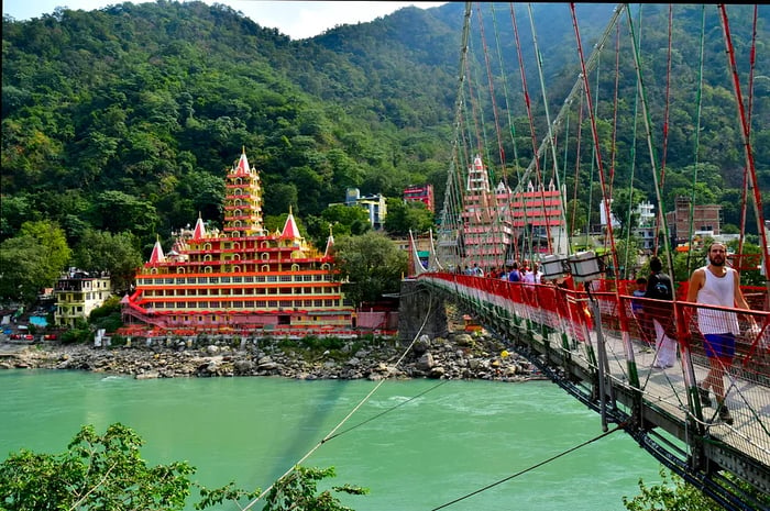 People traverse a suspension bridge over a wide river toward a temple encircled by trees.