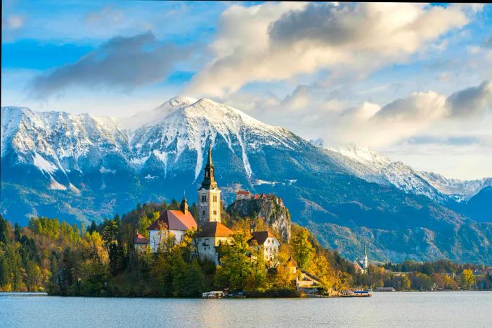 A stunning church emerges from the trees in front of a lake, framed by majestic mountains in the distance.