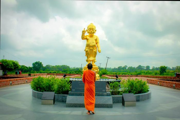 A monk dressed in orange robes stands before a golden Buddha statue