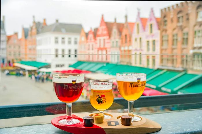 Three beer glasses perched on a ledge with a backdrop of colorful Belgian houses